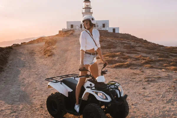 A young woman wearing a helmet sits on a white ATV on a dirt path leading towards a white lighthouse at sunset in Mykonos