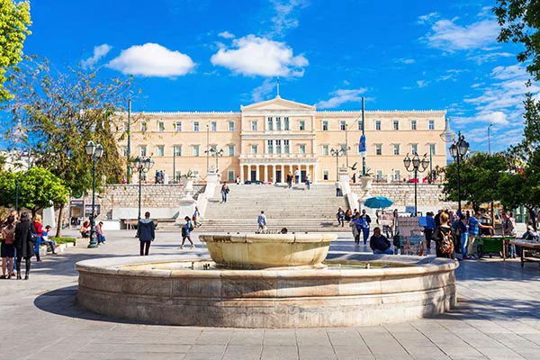 Syntagma squaare with a large fountain in the foreground and the grand Hellenic Parliament building elevated on a platform with steps in the background under a bright sky.