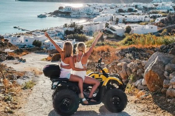 Two people with their arms raised ride a yellow ATV on a dirt path overlooking a coastal town with white buildings and the sea.